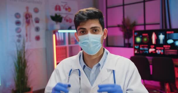 Close up portret sympatyczny pewny siebie męski chemik w mundurze medycznym i maski twarzy, które zakładając jego okulary patrząc w aparat fotograficzny w ciemnym oświetlonym laboratorium — Wideo stockowe