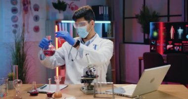Koruyucu maskeli ve üniformalı çekici deneyimli kimyager akşam laboratuarında kimyasal sıvılarla çalışıyor.