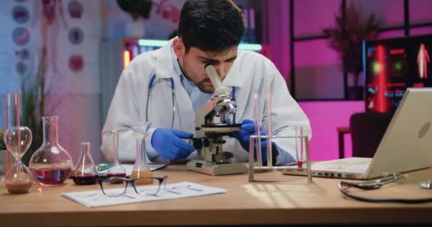 Подходящий трудолюбивый бородатый ученый в белом халате просматривает образцы через микроскоп в вечерней лаборатории и вводит результаты в бумажный отчет, замедленное движение — стоковое видео