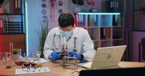Jóképű, magabiztos, tapasztalt fiatal vegyész egyenruhában és orvosi maszkban, mikroszkóp alatt mintákkal dolgozik, és folyadékokat keverget lombikban az esti laborban végzett kísérlet során. — Stock videók