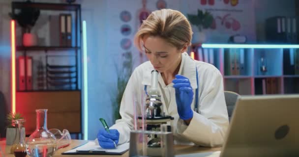 Привлекательная уверенная в себе опытная женщина-химик в белом халате и стерильных перчатках, отмечающая важные детали в отчете при просмотре проб под микроскопом — стоковое видео