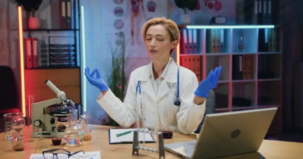 Beautiful overworked high-skilled blond woman doctor meditating with closed eyes and mudra hands in modern medical workroom in the evening — Stock Video