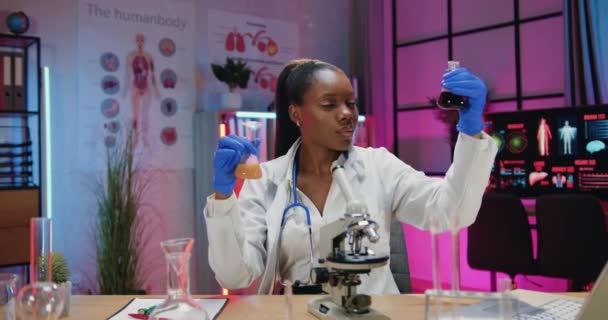 Leuke positieve gekwalificeerde slimme Afrikaanse Amerikaanse vrouwelijke wetenschapper in uniform en beschermende handschoenen zitten aan het bureau in 's avonds lab en werken met flessen met chemische vloeistoffen — Stockvideo