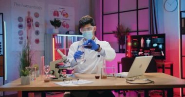 Üniformalı ve koruyucu maskeli yetenekli çekici erkek kimyager akşam laboratuarında sıvılarla kimyasal deneyler yapıyor.