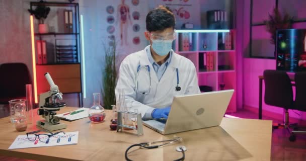 Привлекательный концентрированный трудолюбивый ученый в защитной маске, работающий на ноутбуке во время сверхурочной работы в вечерней лаборатории — стоковое видео