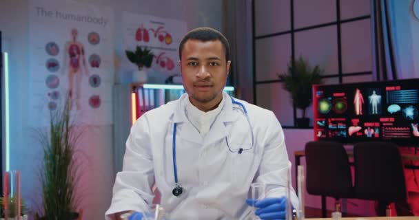 Handsome confident qualfied young african american chemist in white coat posing on camera with glass flask and test tubes filling with fluids in evening lab — Stock Video