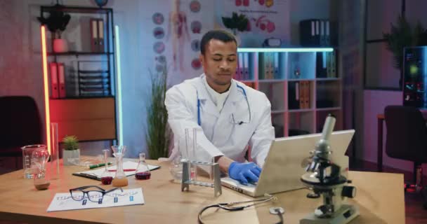 Goed uitziende zelfverzekerde doelbewuste qulified Afro-Amerikaanse mannelijke chemicus in uniform werken aan laptop en het bekijken van chemische oplossingen in kolven tijdens zijn werk in 's avonds lab — Stockvideo