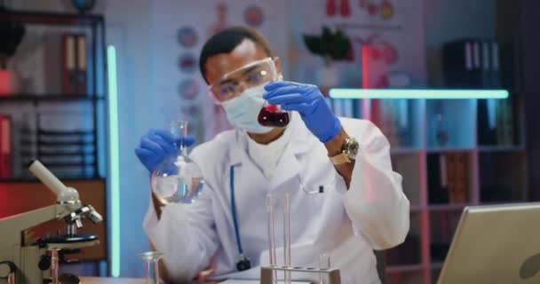 Likable confident purposeful qualified african american male chemist in protective mask,eyewear ,gloves and white coat conducting chemical experiment with fluids in dark lighted lab — Stock Video