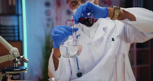 Verantwoordelijke Afrikaans-Amerikaanse mannelijke chemicus met beschermend masker, handschoenen, brillen en witte vacht die chemisch experimenteert met oplossingen en kolven — Stockvideo
