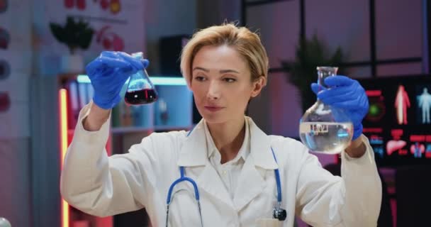 漂亮而自信的金发女化学家，身穿制服，戴着手套，在晚上的实验室里对玻璃瓶进行化学研究，并以正面积极的表情看着相机 — 图库视频影像