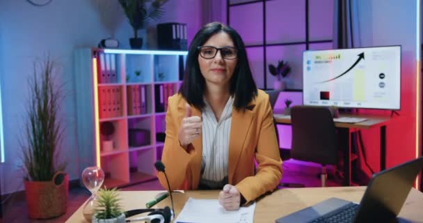 Buena apariencia positiva hábil mujer de negocios de cabello oscuro en gafas y ropa formal mirando a la cámara en la oficina de la noche y haciendo gestos pulgares hacia arriba — Vídeo de stock