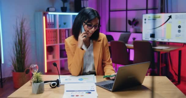 Обаятельная успешная опытная темноволосая женщина-офисный менеджер в стильной одежде и очках, работающая на ноутбуке и разговаривающая по телефону в вечернем офисе — стоковое видео