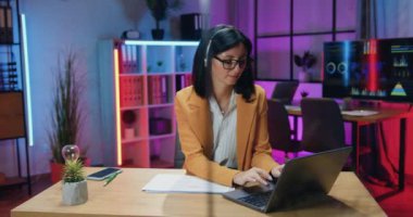 Oldukça kendine güvenen, kulaklıklı, şık giyinen, gözlüklü, laptopta çalışan ve akşam ofisinde dostça bir yüz ifadesiyle kameraya bakan modern, deneyimli bir esmer.