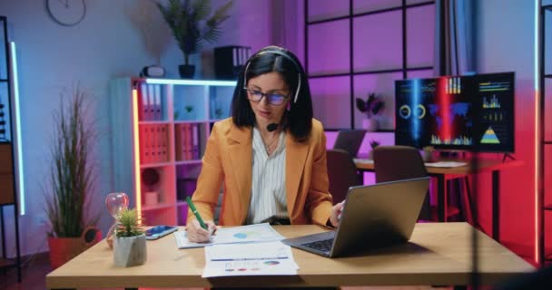 同僚とオンラインチャット中にコンピュータの前に座ってヘッドセットとメガネで魅力的な正の成功した熟練したビジネス女性がビジネスコンセプト — ストック動画