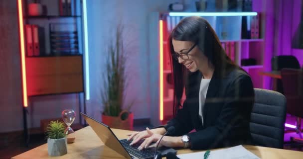 Красивая улыбающаяся успешная опытная брюнетка в стильной одежде сидит на рабочем месте в вечернем офисе и печатает на ноутбуке, бизнес-концепция — стоковое видео