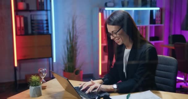 Boa aparência sorrindo bem sucedido experiente morena no desgaste elegante sentado em seu local de trabalho no escritório da noite e digitando no laptop, conceito de negócio — Vídeo de Stock