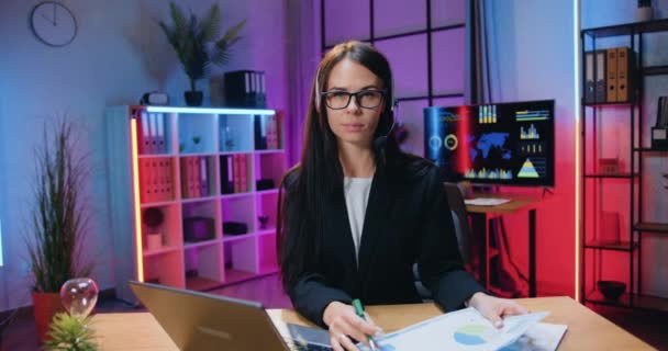 Hermosa confianza moderna elegante experimentada joven morena en auriculares y gafas posando en la cámara en la sala de trabajo iluminada por la noche, retrato de negocios — Vídeos de Stock