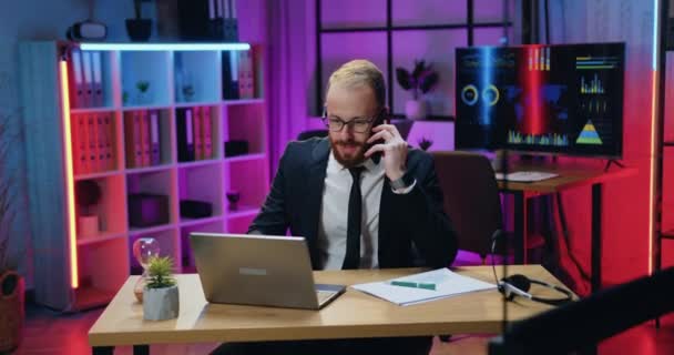 Workflow w biurze, gdzie przystojny zajęty celowo wykwalifikowany młody brodaty biznesmen w formalnym stroju w okularach rozmawia przez telefon i przegląda raporty papierowe i informacje komputerowe — Wideo stockowe