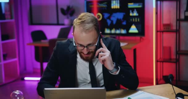 Aantrekkelijk positieve succesvolle ervaren bebaarde zakenman die telefoneert met collega of klant en 's avonds naar het laptopscherm kijkt — Stockvideo