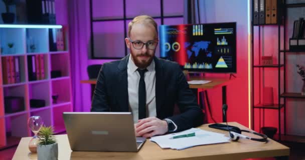 Szerethető pozitív magabiztos szakképzett szakállas üzletember formális viselet szemüvegben ül a kamera előtt az esti irodában, és barátságos arckifejezéssel — Stock videók
