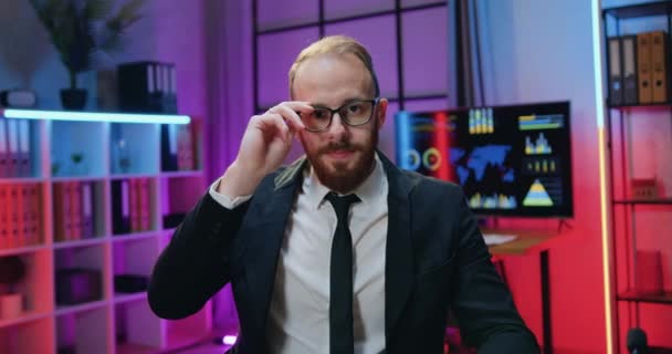 Close-up portret van knappe zelfverzekerde vriendelijke gekwalificeerde bebaarde zakenman in donker pak die het nemen van zijn bril terwijl het kijken in de camera in het avondkantoor — Stockvideo