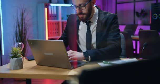 Sympathisch selbstbewusster, fleißiger, bärtiger Geschäftsmann in formeller Kleidung und Brille, der an seinem Arbeitsplatz sitzt und spät im Büro am Computer arbeitet — Stockvideo