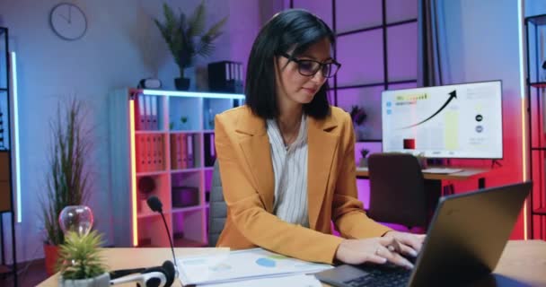 Menarik positif tegas brunette terampil dalam gaya memakai dan kacamata duduk di depan komputer dan bekerja dengan informasi pada layar dan laporan keuangan di kantor malam — Stok Video
