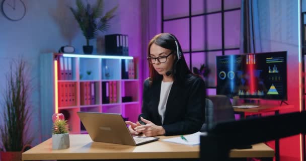 Concepto de negocio donde hermosa confiada hábil elegante mujer de negocios de pelo oscuro en auriculares celebración de reunión en línea con sus subordinados en el ordenador portátil de la oficina de la noche — Vídeo de stock