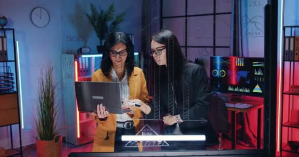 Abbastanza fiducioso operoso altamente qualificato due donne d'affari che lavorano insieme vicino al muro di vetro in ufficio sera, registrano segni importanti dal rapporto del computer — Video Stock