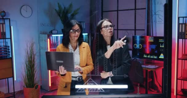 Pretty confident industrious high-skilled two businesswomen working together near glass wall in evening office,they recording important marks from computer report — Stock Video