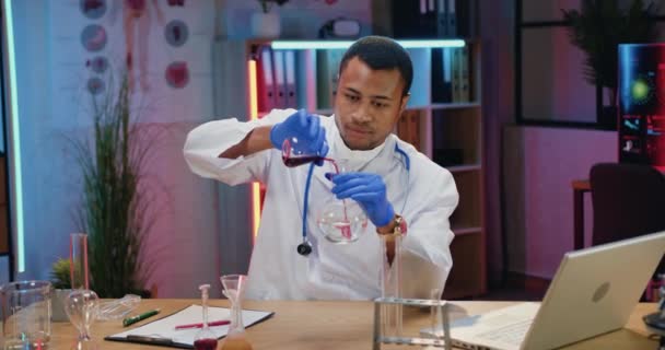 Jóképű koncentrált, keményen dolgozó afro-amerikai férfi vegyész fehér köpenyben kémiai kísérleteket végez folyadékokkal az esti laborban. — Stock videók