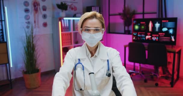 Laboratuvar önlüklü, koruyucu gözlüklü, maske takan, kameranın önünde oturan ve elinde sıvı olan iki şişe tutan güzel ve kararlı sarışın kadın kimyager. — Stok video