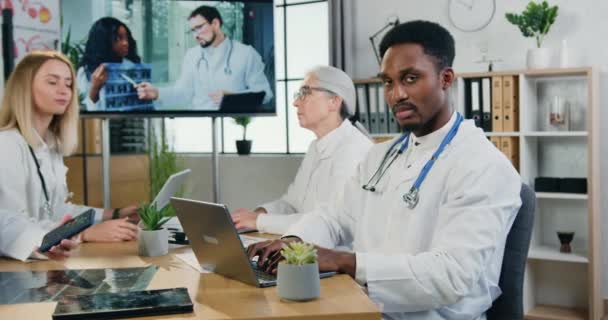 Workflow in clinic where attractive serious professional african american doctor looking into camera during joint meeting with anothers members of medical team — Stock Video