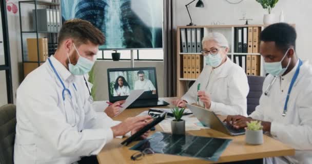 Attrayant hautement qualifiés confiants médecins multiethniques masculins et féminins sous masques travaillant ensemble dans le bureau médical lors d'une séance d'information en ligne avec des collègues via ordinateur, concept covid-19 — Video