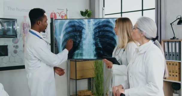 Доброзичливі кваліфіковані багатоетнічні лікарі, які обговорюють результати рентгенівського випромінювання легень, зафіксованого на бічній дошці в клінічному кабінеті під час спільної ради, концепція медицини — стокове відео