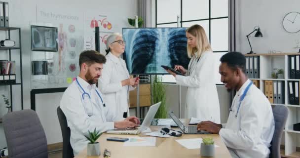 Concepto de medicina donde atractivos médicos multiétnicos experimentados y seguros trabajan juntos en el consultorio médico, hombres escribiendo en computadoras portátiles mientras las mujeres discuten la radiografía de tórax y pulmones en pantalla — Vídeo de stock