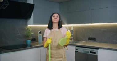 Profesyonel temizlikçi kadının portresi. Plastik koruyucu sarı eldivenler ve önlük takıyor. Elinde paspas ve sprey deterjan tutuyor..