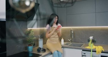 Önlük giymiş, mutfak yüzeyinde oturan genç bayan ev mutfağını temizledikten sonra akıllı telefon kullanıyor..