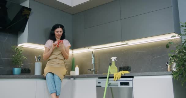 Belle femme au foyer caucasienne habillée en tablier assis sur la table tenant gadget moderne dans les mains et écouteurs pendant le nettoyage de la maison à la cuisine à la maison — Video