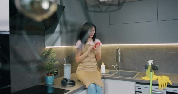 Jovem dona de casa vestida com avental sentado na superfície da cozinha usar smartphone após a limpeza em casa cozinha. — Vídeo de Stock
