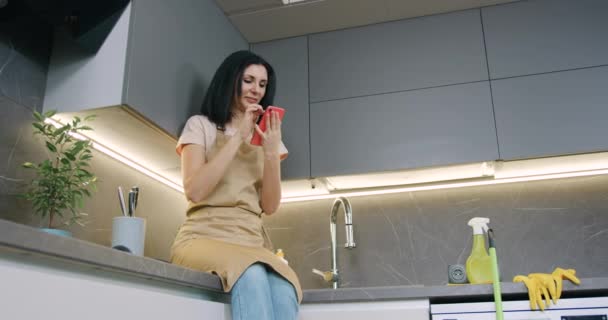 Όμορφη μελαχρινή γυναίκα στην κουζίνα ποδιά με smartphone στα χέρια συγκρατημένο γέλιο, ενώ κάθεται στην κουζίνα στο σπίτι — Αρχείο Βίντεο