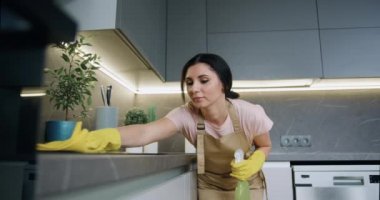 Sarı lastik eldivenli genç ev hanımı mutfak tezgahındaki elektrikli sobayı deterjanlı ıslak sarı bir bezle yıkıyor, spreyliyor ve siliyor. Ev mutfağı temizlik konsepti