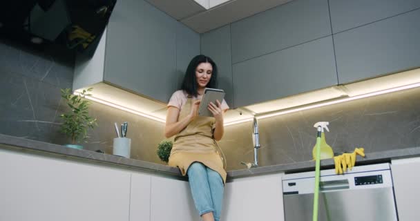 Bela mulher morena caucasiana pensativa com o uso de computador tablet de navegação, durante a limpeza em casa de cozinha — Vídeo de Stock