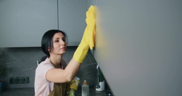 Vue latérale de la jeune femme caucasienne sérieuse dans un tablier et des gants de protection pendant le nettoyage de la surface de la cuisine à la maison — Video