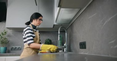 Evdeki modern mutfakta, sarı eldivenli, beyaz tabak yıkayan genç bir kadın.