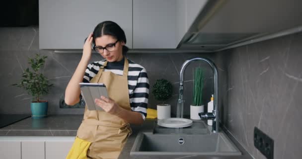 Молодая женщина в очках смотрит на экран планшета во время мытья посуды на кухне дома, рутина, образ жизни — стоковое видео