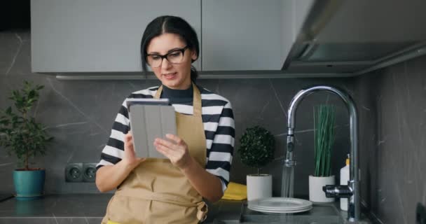 Portrait de jeune belle femme dans les lunettes à l'aide de l'écran de tablette numérique lors du lavage de la vaisselle à la maison de la cuisine, routine — Video