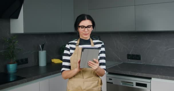 Орієнтована красива і усміхнена біла жінка в кухонному фартусі використовує планшетний гаджет, фрілансер, який працює з дому на домашній кухні — стокове відео