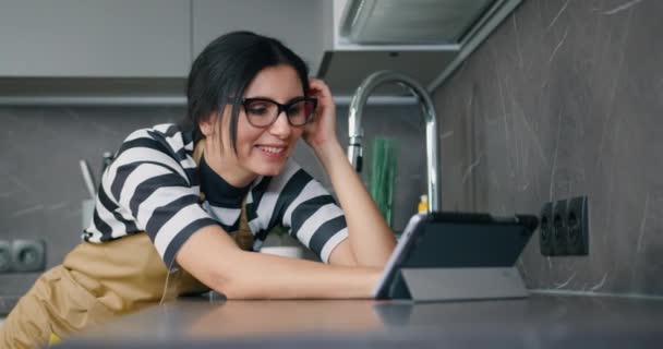 Молодая кавказская улыбающаяся женщина в очках с помощью интернета на планшетном устройстве во время домашней рутины на домашней кухне — стоковое видео