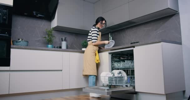 Bela dona de casa morena caucasiana em avental em pé na frente da máquina de lavar louça e colocando pratos de lavagem. Interior da cozinha. — Vídeo de Stock